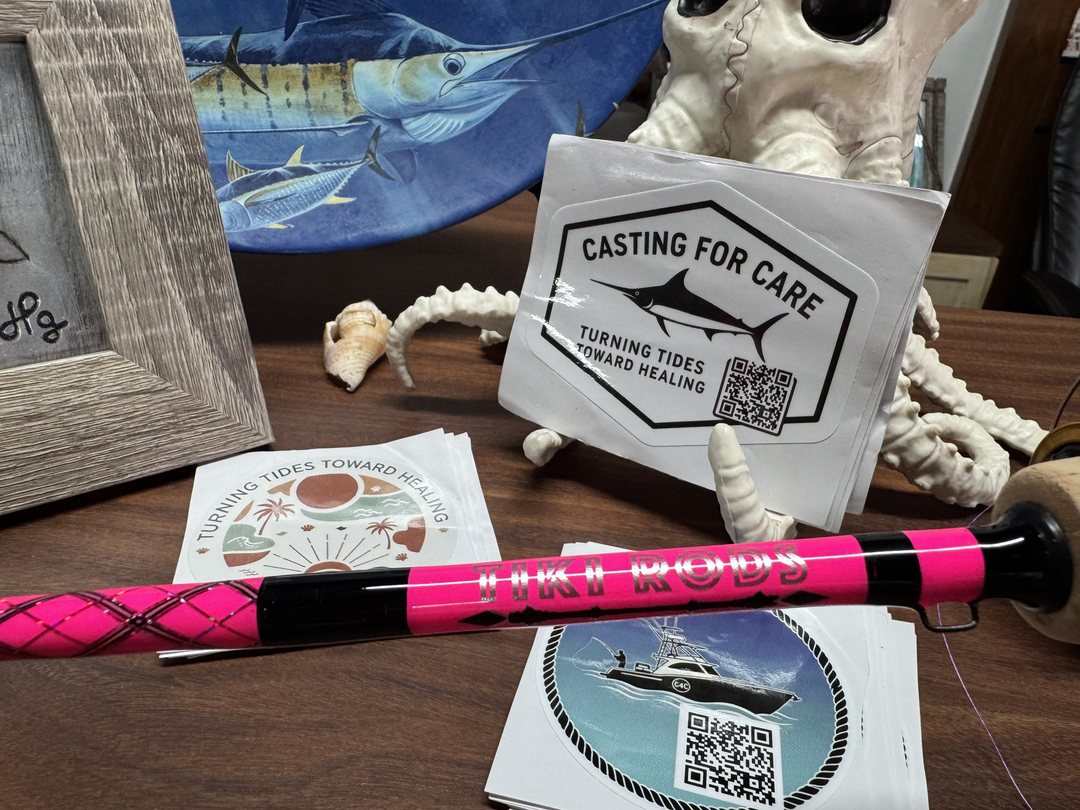 A pink and black Tiki Rods blank laid beside Casting for Care stickers reading "turning tides toward healing", on a desk in the shop