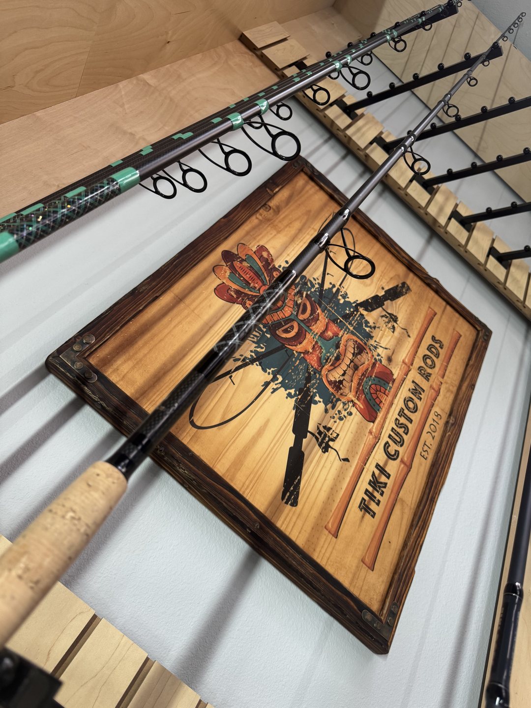 The carved Tiki Custom Rods sign, established 2018, hanging on the shop wall beside rods in the rack