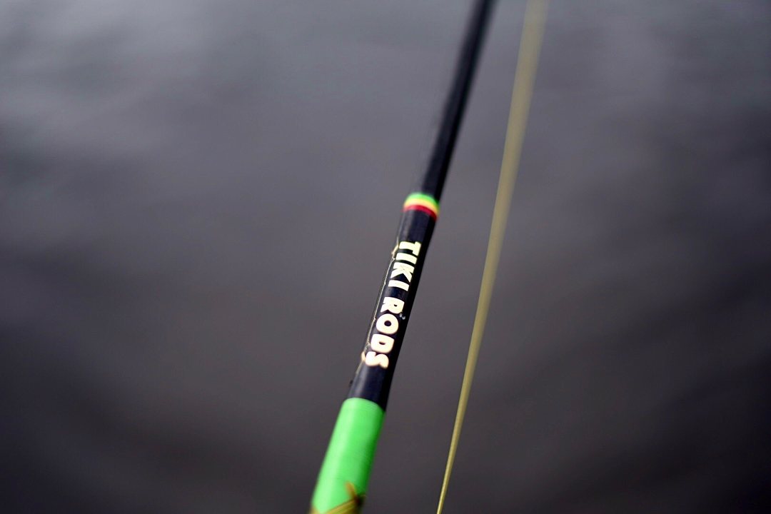 Close-up of the Tiki Rods decal on a blank above a green grip, line crossing the frame and open water behind