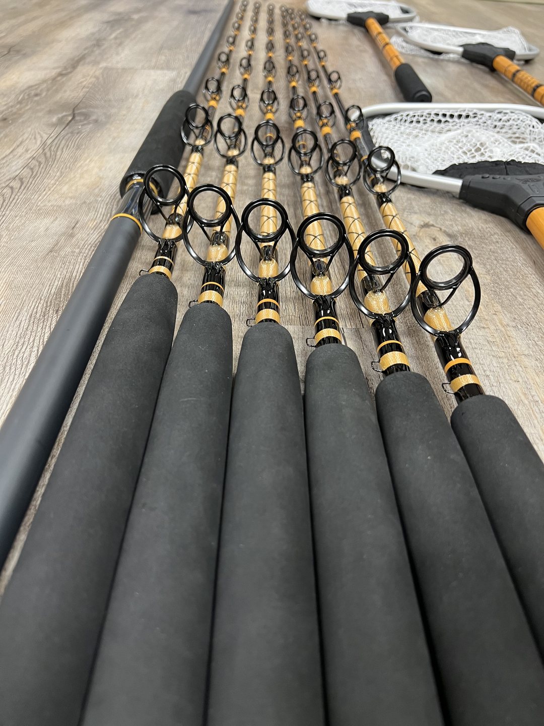 A row of offshore rods with gold and black decorative wraps, oversized guides and foam grips, on a grey wood floor