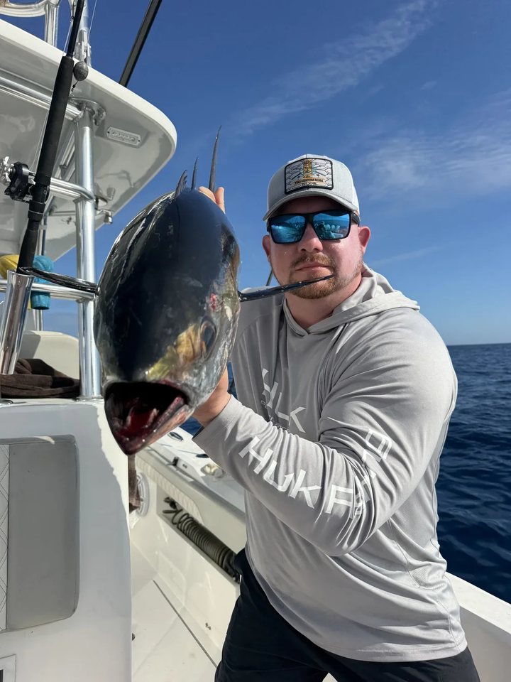 Jack Maglione on a center console offshore, holding up a tuna he has just landed, wearing a Tiki Custom Rods cap and sunglasses with open water behind him