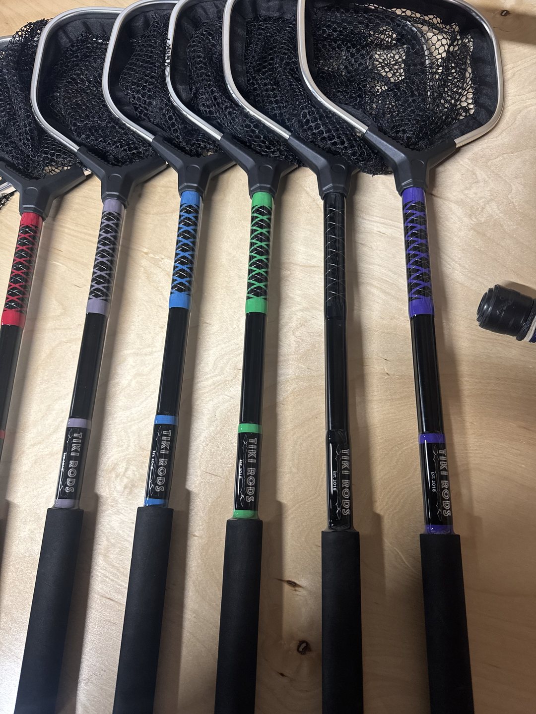 Six landing nets laid on a bench, their handles cross-wrapped in red, grey, blue, green, black and purple