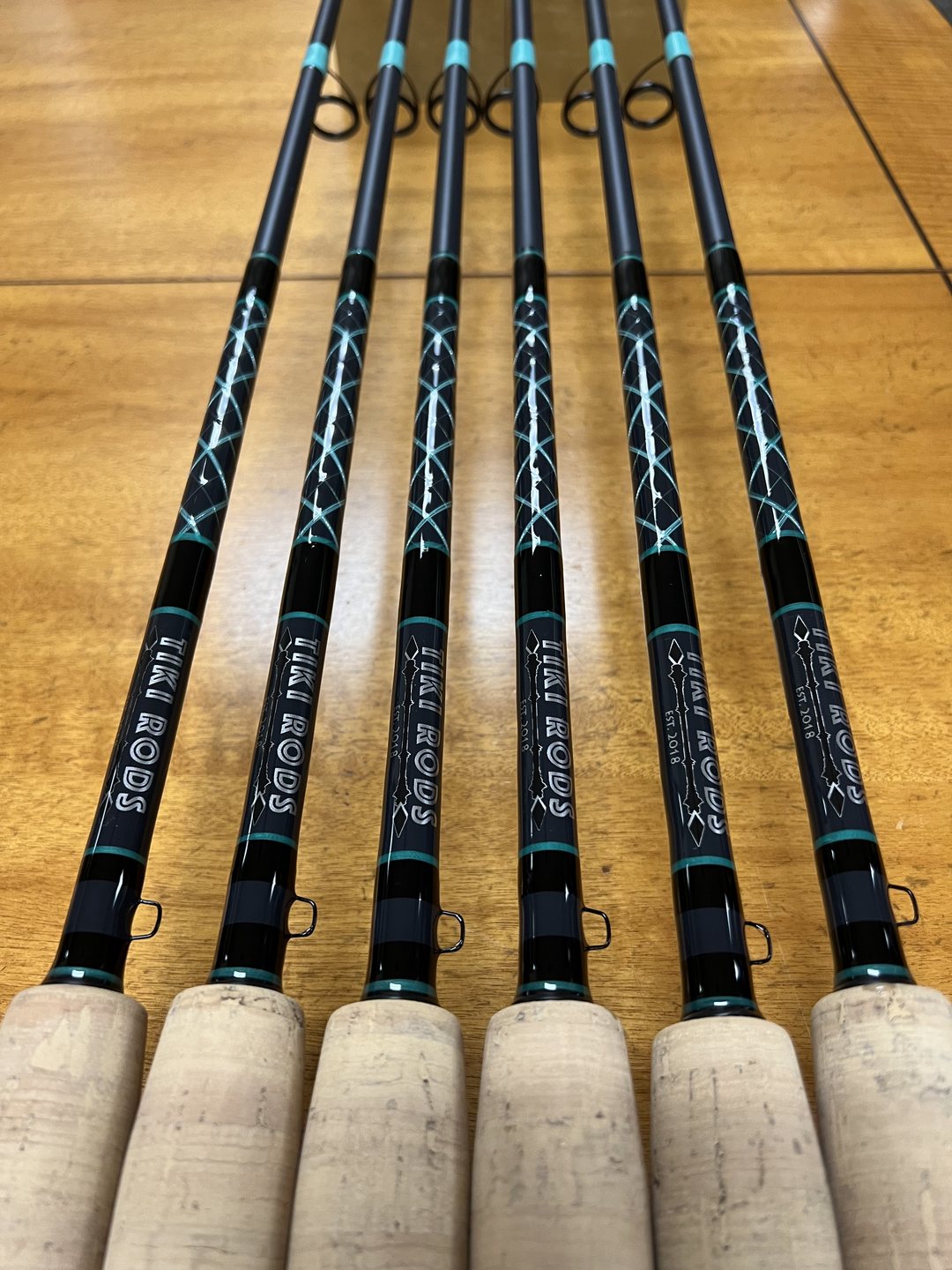 Six rods with teal cross wraps over black blanks and cork grips, each carrying the Tiki Rods decal